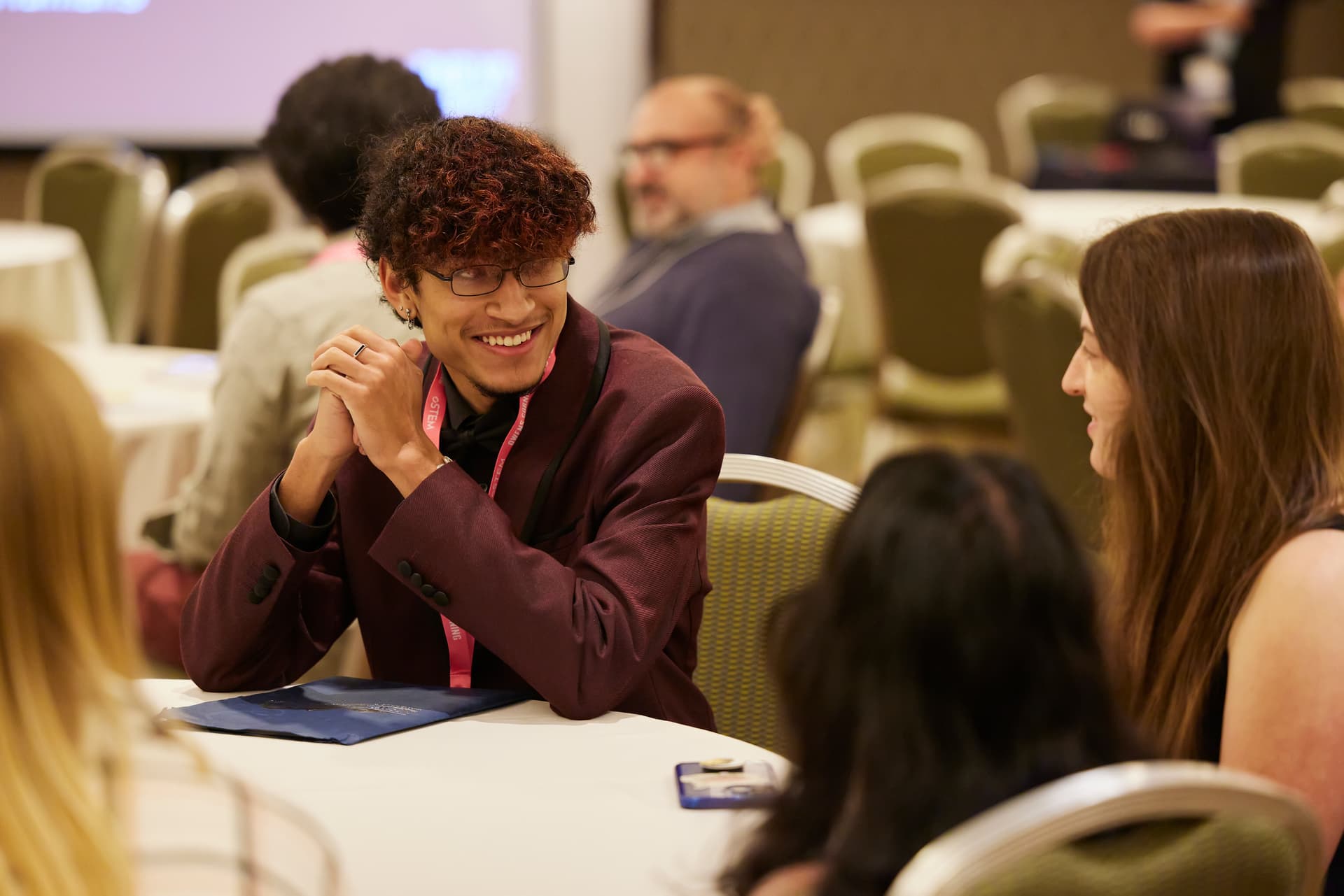 A decorative image of oSTEM conference attendees sitting around a table. One attendee is in focus looking at another attendee, while four other attendees can be seen blurred in the foreground and background.